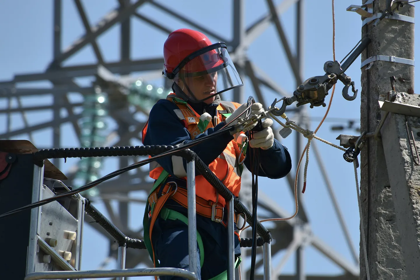Lineworker in a safety harness and face shield adjusting a cable at a utility pole.